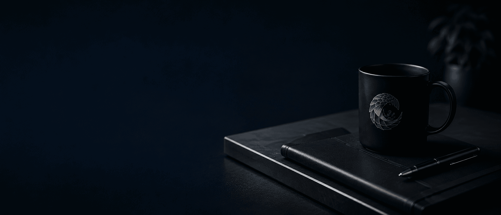 Black LeKoj Studio mug, notebook, and pen on a dark desk with soft lighting and greenery.